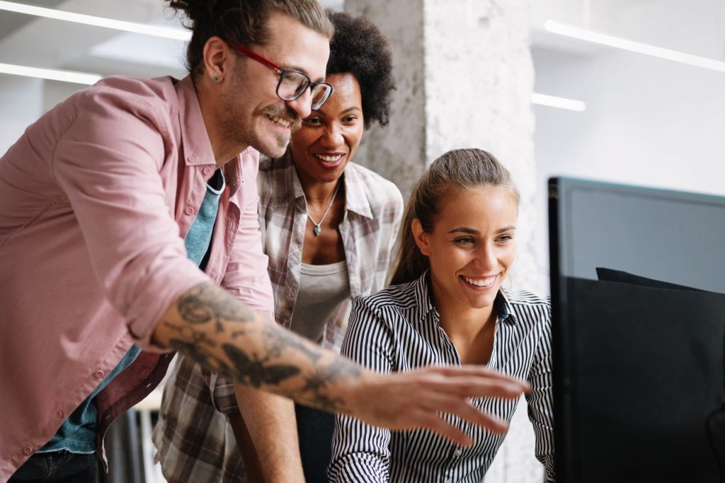 Three people (man and two women) gathered around a computer screen. The man is pointing to the screen.